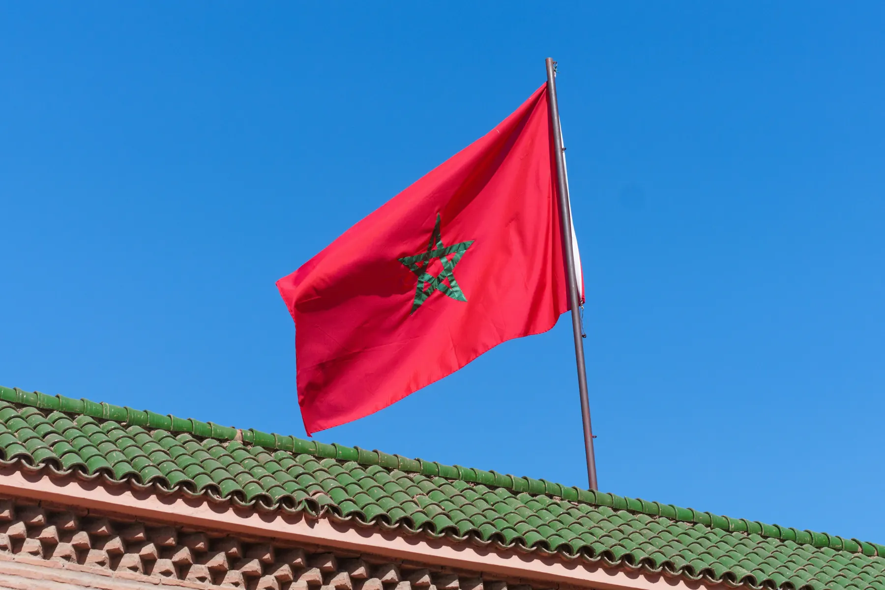 Le drapeau du Maroc, rouge à l'étoile verte, flotte au-dessus d'un toit de tuiles vertes sous un ciel bleu